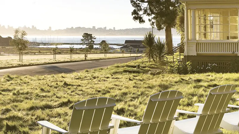 Four white wooden chairs are arranged on a grassy lawn overlooking a tranquil body of water, with a tree and a large house on the right. In the distance, boats, a dock, and a city skyline under a hazy sky evoke the serene ambiance of California spa retreats.