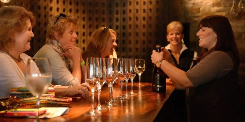 A group of women sits at a wooden bar counter with several empty wine glasses in front of them. A woman on the right side is holding and presenting a wine bottle to the group. The setting appears cozy, with warm lighting and a wall with wine racks in the background—perfect for food and wine tours in Carmel.