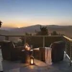 A scenic outdoor patio at one of the best luxury hotels in Monterey Peninsula overlooks a distant sunset beyond rolling hills. The patio features wicker chairs gathered around a small table, a lit lantern, and a candle. A wooden table to the left holds a tray with a drink and another candle, adding to the tranquil atmosphere.