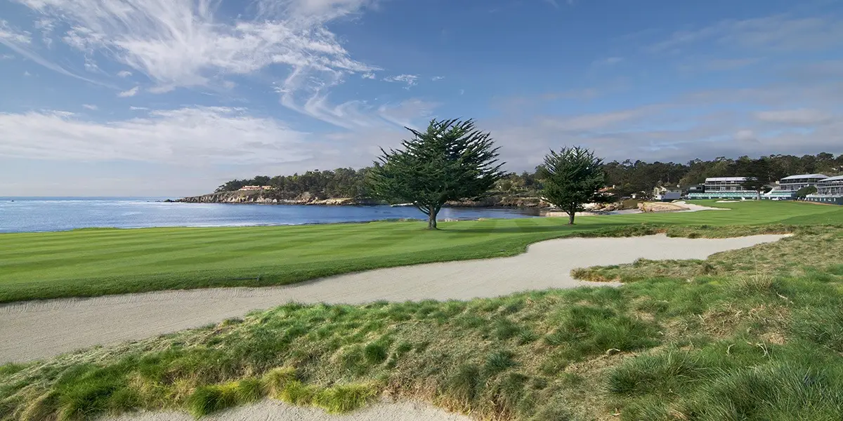 A scenic view of a coastal golf course featuring a green fairway with a sand bunker in the foreground, two tall trees near the center, and a tranquil body of water with a rocky shoreline and buildings in the background. The sky is partly cloudy, making it one of the best golf courses Monterey Peninsula has to offer.