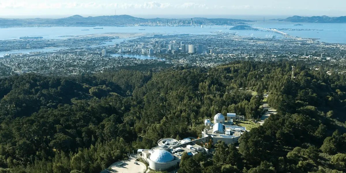 Aerial view of an observatory complex nestled in a forested area with a cityscape and waterfront in the background. Several domed observatory buildings are visible among the trees, inviting visitors to explore East Bay activities while enjoying the sprawling urban landscape and bodies of water in the distance.
