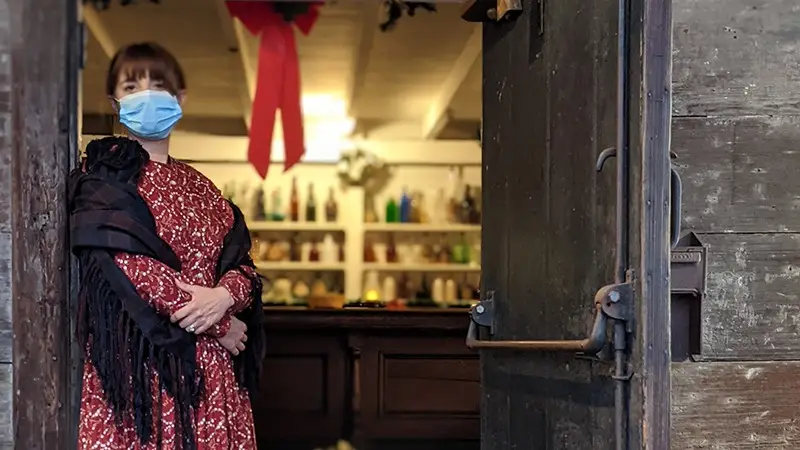 A person wearing a red patterned dress, black shawl, and a blue mask stands in the doorway of an old-fashioned building on the Monterey Peninsula. Behind them, shelves filled with various bottles and jars are visible. A holiday wreath with a large red bow hangs inside the doorway, capturing the festive spirit of December.