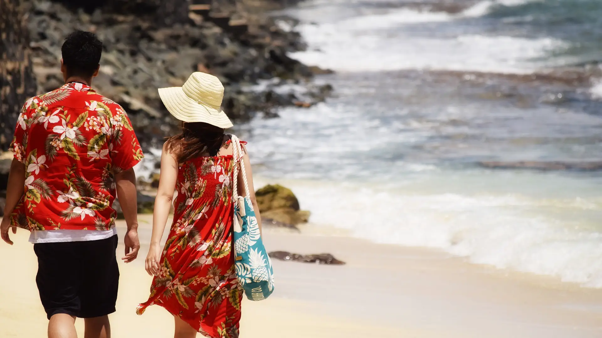 A couple wearing matching red floral outfits walks hand in hand along a sandy beach, embodying the spirit of aloha. The woman has a wide-brimmed hat and a blue tote bag with white patterns, and waves are crashing on the shore nearby, making this scene fit for any list of top 15 Hawaiian words to know.