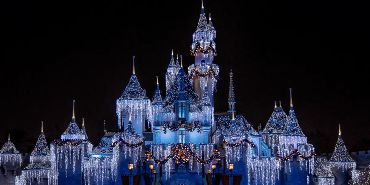 A grand castle illuminated at night with an array of sparkling lights. Among the coolest things at Disneyland during holidays, it features multiple towers and spires adorned with festive decorations including wreaths, garlands, and icicle-like lights, creating a magical winter wonderland atmosphere.