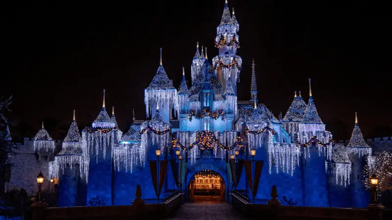 A majestic castle is decorated with festive holiday lights and ornaments. The structure, celebrating Disneyland's 100th anniversary, is adorned with sparkling icicles and wreaths glowing against a dark night sky. The entrance is warmly lit, creating a magical and inviting atmosphere.