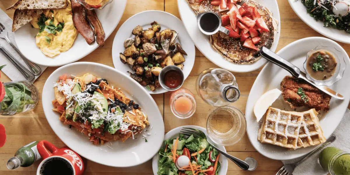 A wooden table is filled with an assortment of breakfast and brunch dishes, including scrambled eggs with bacon, pancakes with strawberries, roasted potatoes, a salad, waffles with fried chicken, and a vegetable-loaded omelette, along with drinks and condiments.