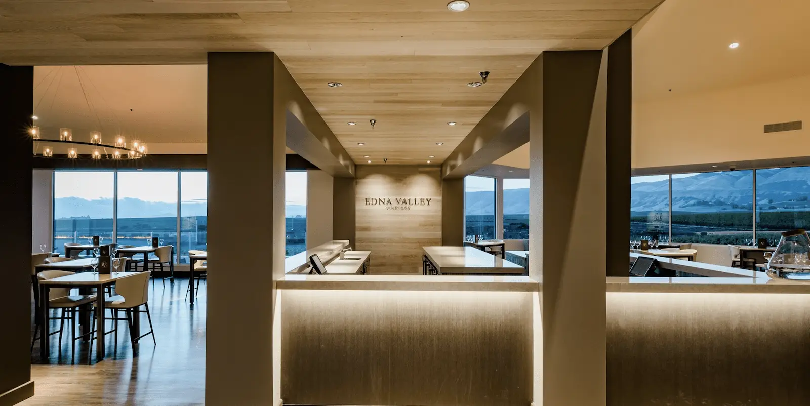 A modern, spacious dining area features large windows with sweeping mountain views. Light wooden ceilings, sleek furnishings, and ambient lighting create an inviting atmosphere. Tables and chairs are neatly arranged, and a sign reads "Edna Valley Vineyard," the perfect spot to taste wine in SLO.