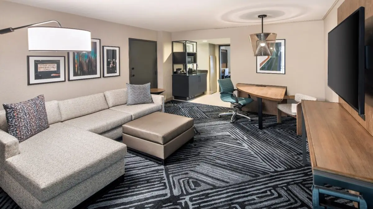 A modern hotel suite featuring a beige sectional sofa with patterned pillows, an ottoman, a large flat-screen TV, a small dining table with a green chair, artistic wall decor, and a kitchenette with a microwave and mini-fridge. It's one of the best value hotels in North Bay.