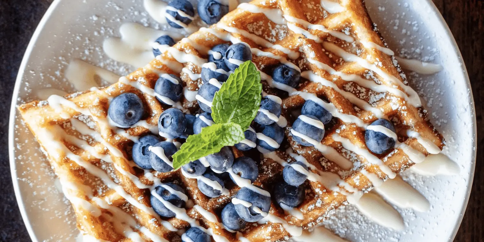 A close-up of a waffle crowned with fresh blueberries, a drizzle of white icing, and a sprinkle of powdered sugar. A mint sprig graces the center for garnish. Served on a white plate, this delectable dish is truly the best breakfast North Bay can offer.