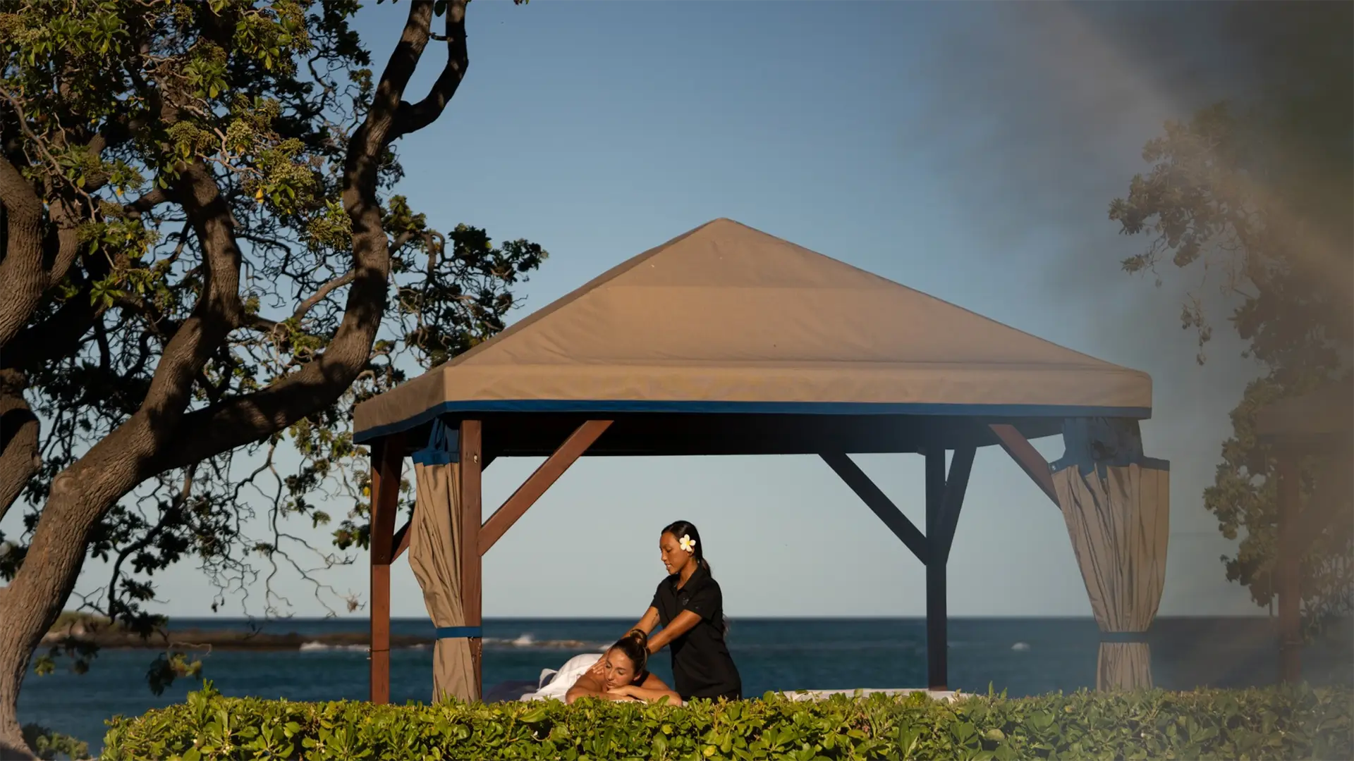 A person is receiving a massage under a beige gazebo by the ocean at a luxurious hotel spa. There are trees on the left side, and the sky is clear with a light haze. The massage therapist is standing, and greenery surrounds the gazebo. This serene escape is one of Big Island's finest offerings.