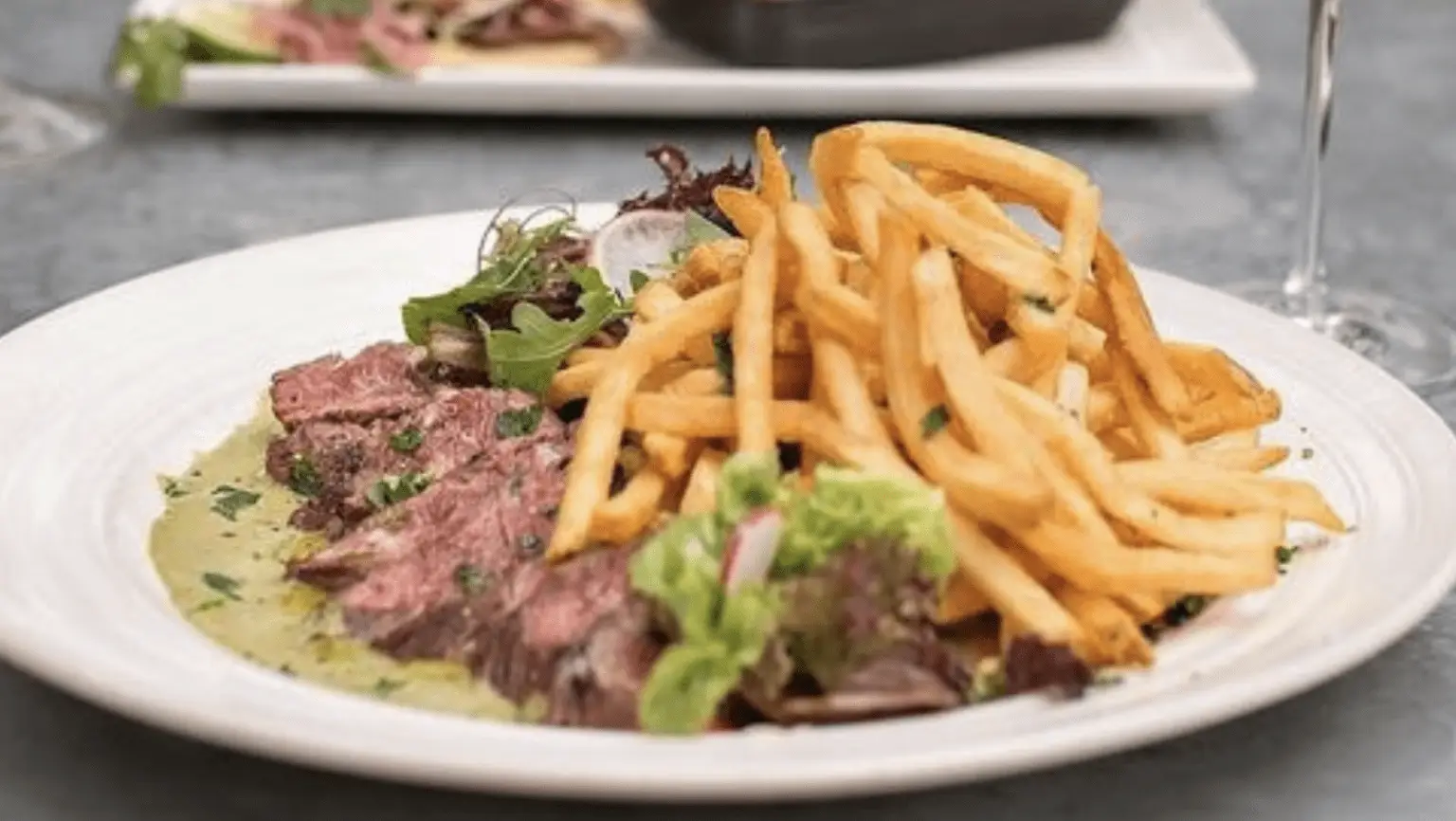 A white plate holds a serving of sliced steak drizzled with a green herb sauce, accompanied by a generous portion of golden French fries and a small side salad with mixed greens. This could be the best dinner in Napa Valley. Another dish is blurred in the background on the table.