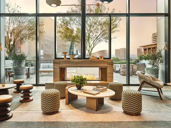 A contemporary lounge with floor-to-ceiling windows showcasing an outdoor terrace. The space features a central fireplace, stylish round ottomans, a low coffee table with books, and modern wooden furniture. Potted plants and city views can be seen outside in this eco-friendly haven typical of California Eco hotels.