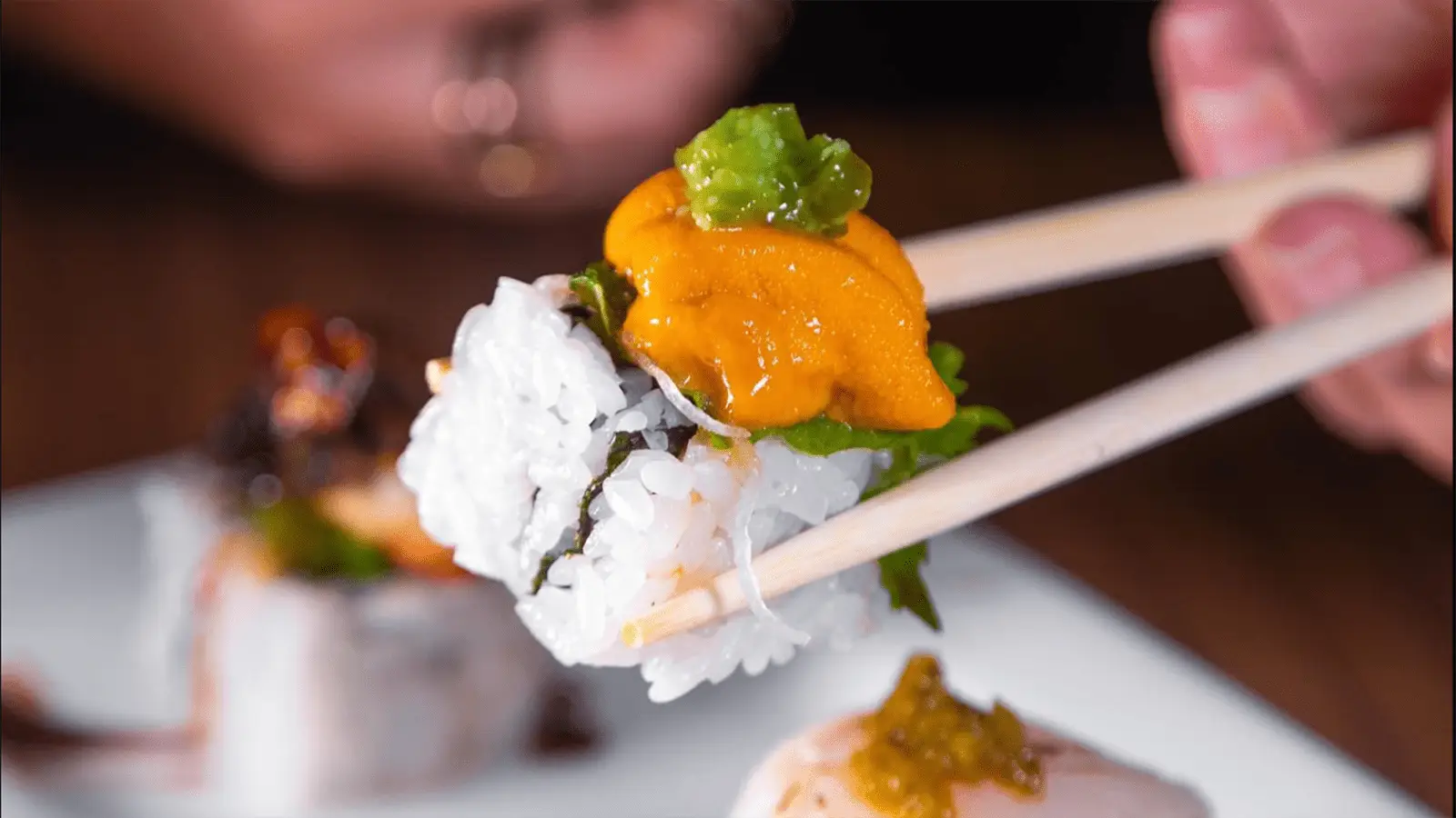 Close-up of chopsticks holding a sushi roll topped with vibrant orange sea urchin and green garnish. In the background, another sushi roll sits on a white plate. A hand is slightly blurred, indicating motion. The scene is set on a wooden table at the best sushi spot in Waikiki.
