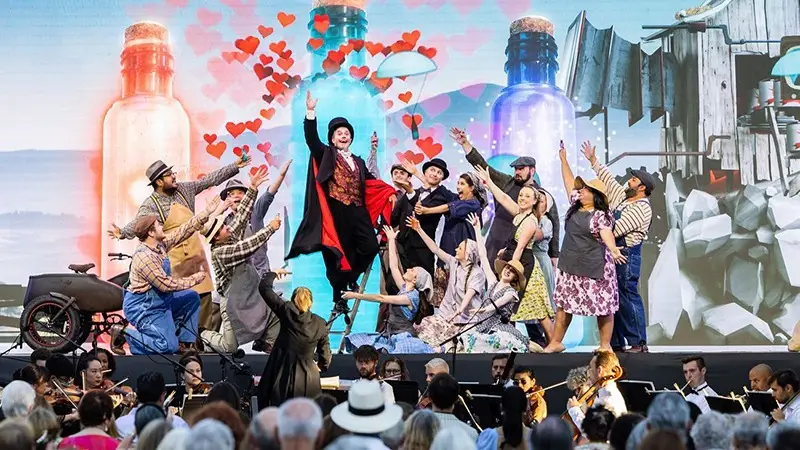 A vibrant outdoor stage scene depicts a lively theatrical performance with actors in colorful costumes. The central figure in a red and black outfit stands out, surrounded by animated cast members and a backdrop featuring large bottles and floating hearts—an enchanting experience, among the best things to do in Wine Country this January.