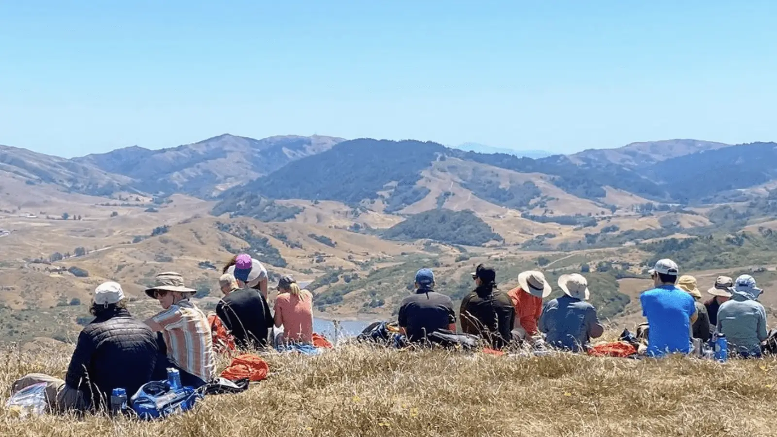 A group of people, dressed casually and wearing hats, sit on a grassy hilltop, facing away from the camera. They overlook a vast landscape of rolling hills and valleys under a clear blue sky. With such scenic beauty and calm surroundings, it's easy to find things to do in North Bay this August.