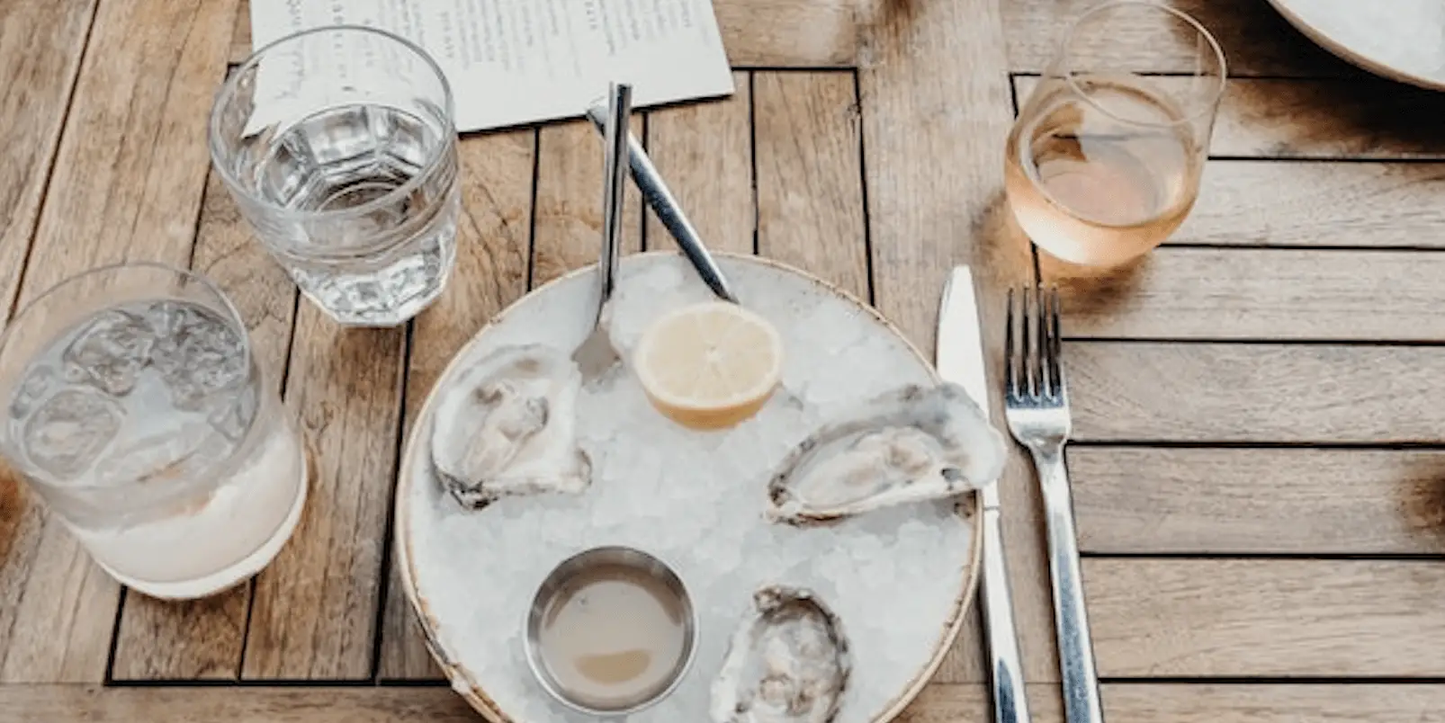 A wooden table holds a plate with ice, four oyster shells, a lemon wedge, and a small dipping sauce bowl. Two glasses, one with a drink and ice and the other with a light-colored beverage, are alongside a menu, fork, and knife on the table—perfect for Happy Hour dining.