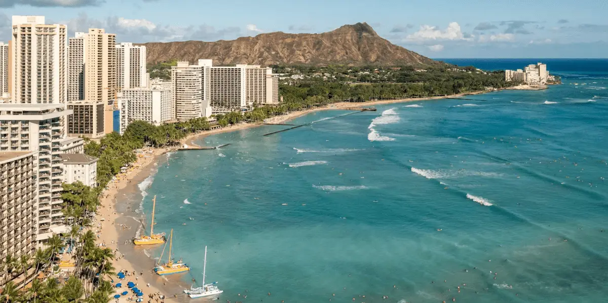 A scenic coastal view featuring a series of high-rise buildings adjacent to a sandy beach with turquoise waters, capturing the essence of statehood pride. Boats and sailboats float near the shore, while a distinctive mountain range rises in the background under a clear blue sky.