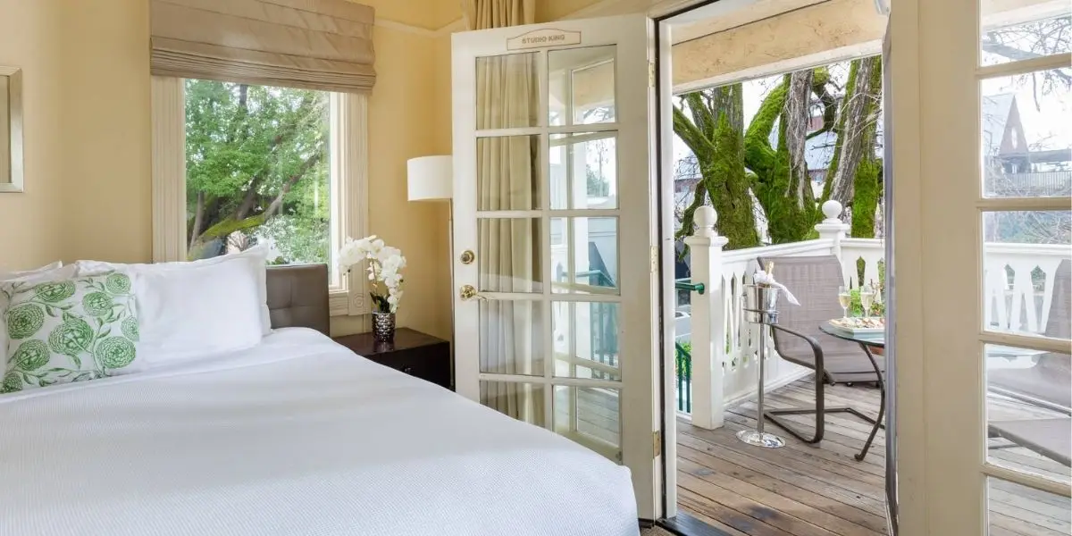 A cozy bedroom with yellow walls features a neatly made bed with white linens and green patterned pillows. French doors open to a wooden balcony with a table and chairs, overlooking lush green trees and a serene outdoor view. White railings adorn the balcony, typical of the best value hotels in Sonoma.