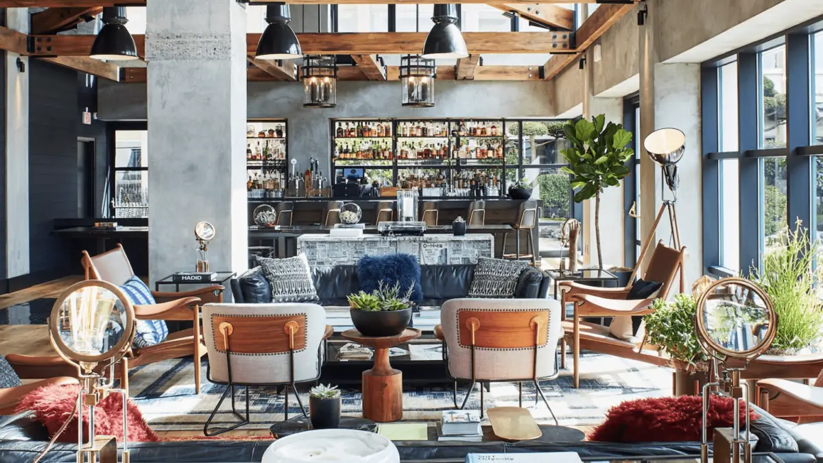 A modern lounge with an industrial design, featuring exposed beams, concrete pillars, and large windows. The space is furnished with leather sofas, wooden chairs, and a mix of contemporary decor. A well-stocked bar is seen at the back with shelves filled with bottles. It's one of the best hip hotels in the Bay Area.