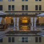 A serene courtyard at one of the best luxury hotels in South Bay, with a reflective pool lined by wooden chairs and tables with umbrellas. The far side features a pergola adorned with white drapes and elegant lanterns, set against a backdrop of a multi-story building with tall windows.