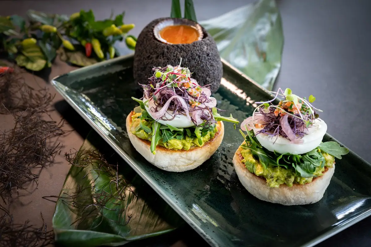 A green rectangular plate holds two pieces of toasted bread topped with avocado, a poached egg, purple sprouts, and microgreens. Beside the toast is a black stone bowl filled with orange sauce. Some greens and small peppers are in the background, making this the best breakfast Kaanapali offers.