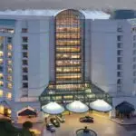 A large Hyatt Regency hotel building is lit up during dusk. The San Jose hotel features multiple floors with numerous windows and a central glass atrium. Cars are parked under a covered entrance, and city lights are visible in the background, making it one of the best value hotels South Bay has to offer.