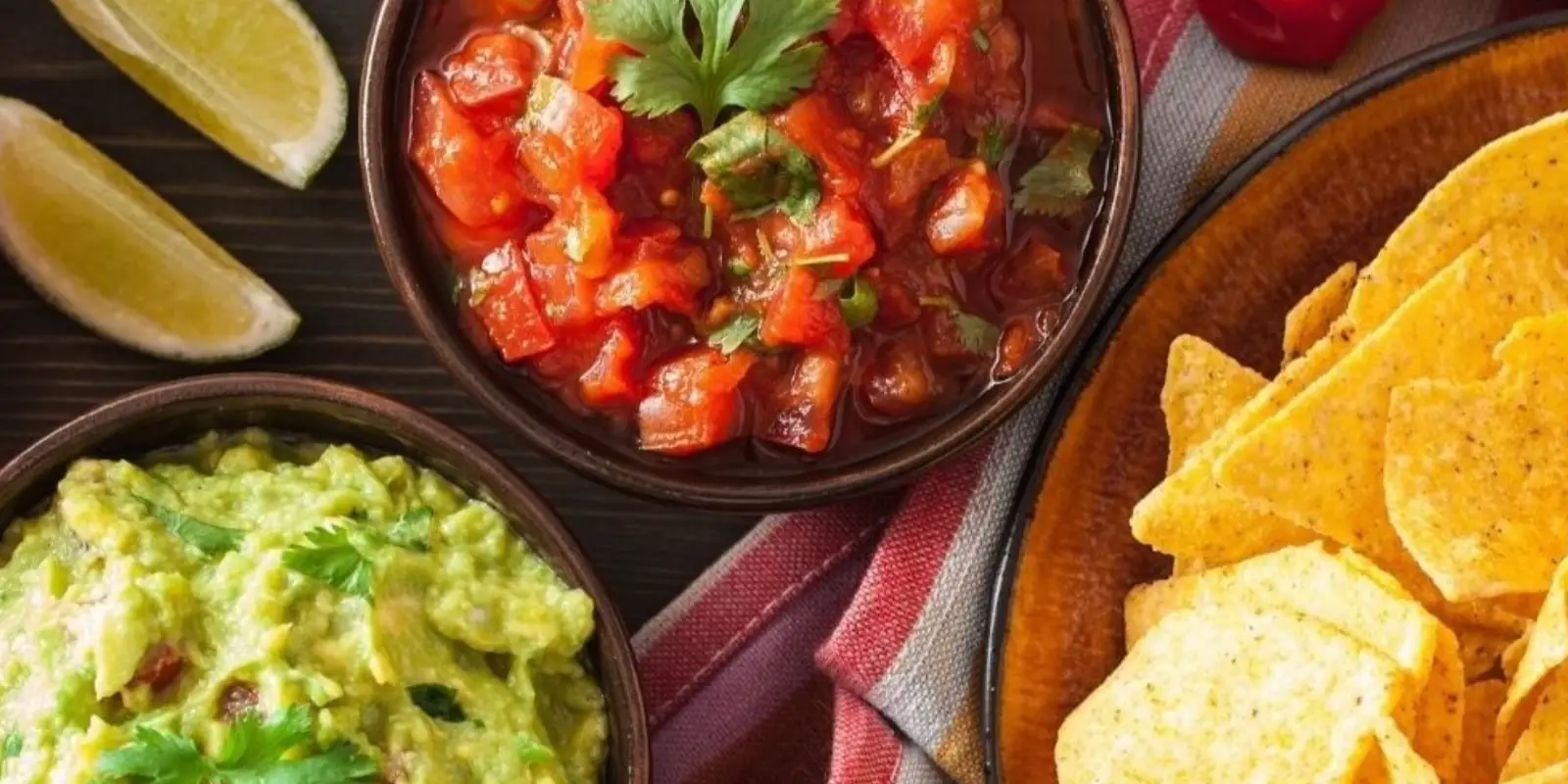 Top view of a table with bowls of guacamole, salsa, and tortilla chips. The guacamole is garnished with cilantro, the salsa has a cilantro garnish, and lime wedges are beside the bowls, all placed on a red and white dish towel—perfect for the best lunch in Tahoe.