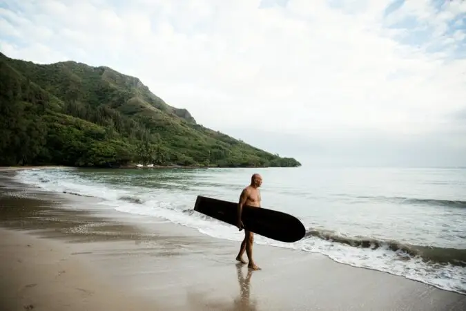 A person walks along Kahana Bay Beach carrying a surfboard under one arm. The beach is bordered by lush green hills and a calm sea. The sky is partly cloudy.