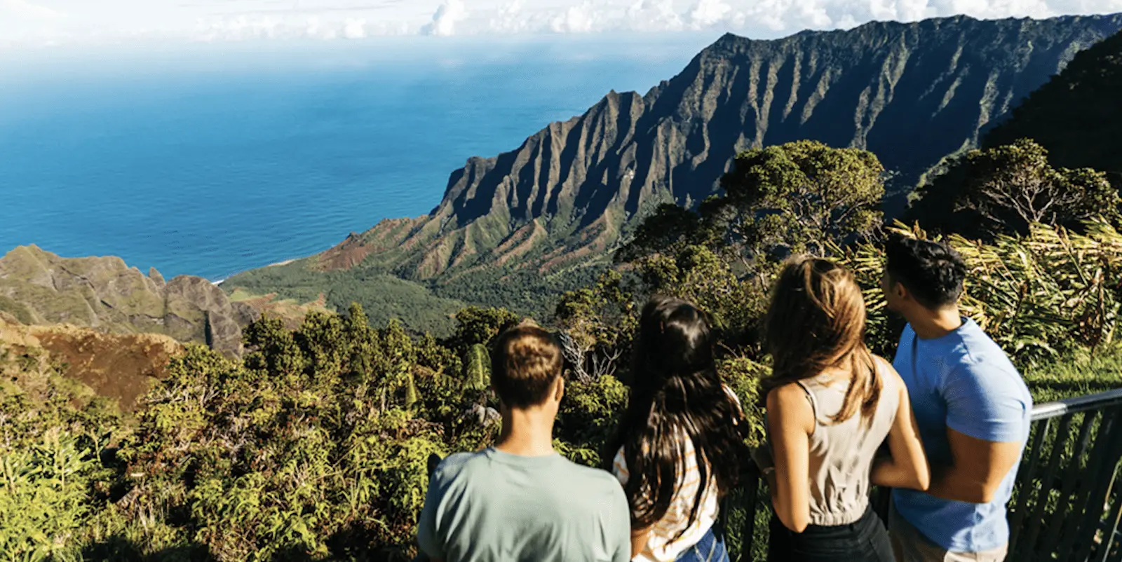 Four people stand at a scenic lookout, viewing lush green valleys and steep cliffs that descend to a blue ocean. The sky is clear with a few clouds, and the group appears to be enjoying the breathtaking landscape—one of the best hikes Kauai has to offer.