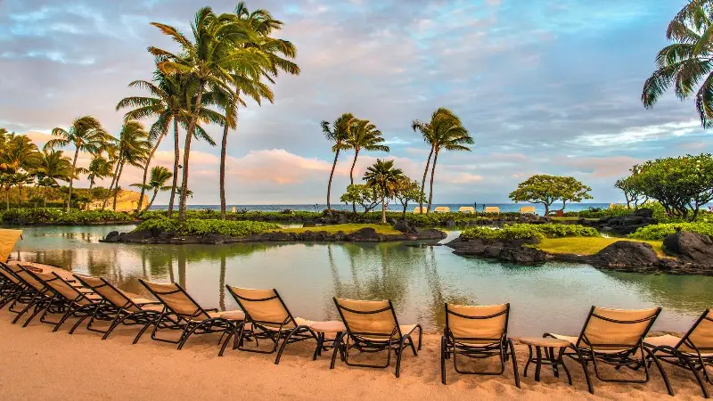 A tropical beach scene featuring a row of empty lounge chairs on a sandy shore, facing a calm lagoon surrounded by rocks and palm trees. The sky is partly cloudy, and the ocean is visible in the background—an idyllic setting often found at the best oceanfront hotels Kauai offers.