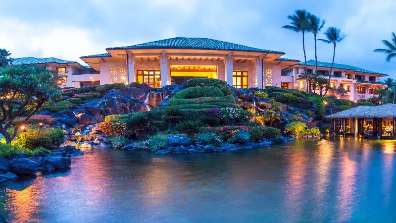 A tropical resort at dusk features a large, well-lit building with a green roof and open architecture. Surrounding it are lush tropical gardens with colorful flowers and palm trees. In the foreground is a serene body of water reflecting the ambient lights, epitomizing one of the best family hotels in Kauai.