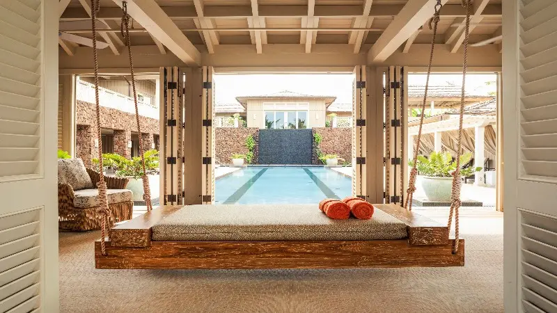 A serene, open-air patio at one of the best spa hotels in Kauai showcases a hanging daybed with orange towels on top, overlooking an inviting pool that stretches towards a modern building in the background. Tropical plants and sleek architecture enhance the tranquil ambiance.