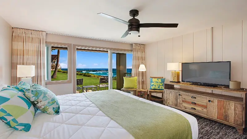 A cozy bedroom featuring a bed with patterned pillows and a green throw blanket, facing a wooden dresser with a large TV. The room has light-colored curtains, a ceiling fan, and a sliding glass door leading to a patio with ocean views and outdoor seating—a perfect example of the best value hotels in Kauai.