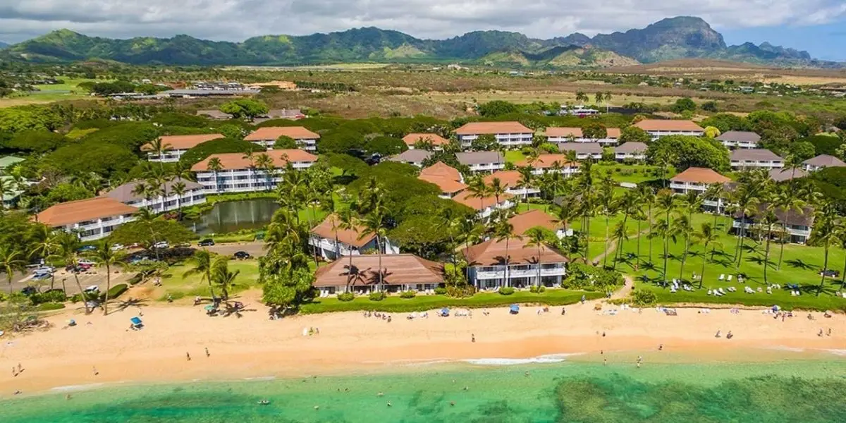 Aerial view of a tropical beach resort with numerous red-roofed buildings surrounded by lush greenery and palm trees. The sandy beach, perfect for families with kids on Kauai, meets clear turquoise waters, backed by mountainous terrain under a partly cloudy sky.