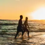 Two people walk hand in hand along the beach at sunset. The sun casts a golden glow over the ocean waves, silhouetting the pair as they stroll by the water near one of the best romantic hotels in Kauai. Palm trees and a distant shoreline are visible in the background.