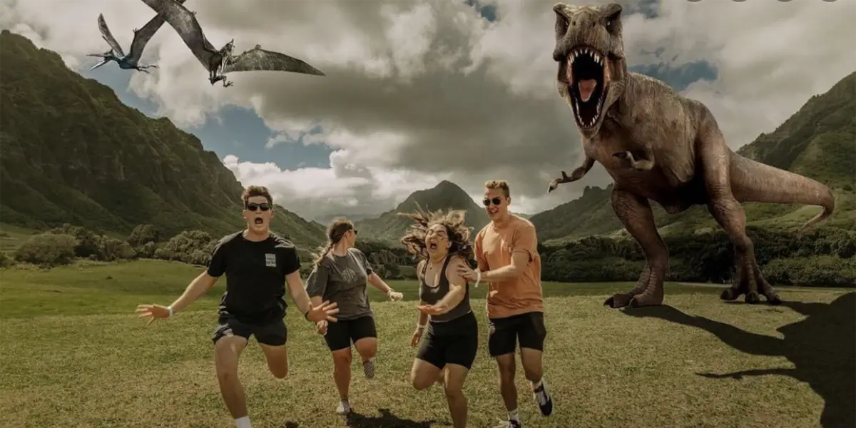 Four people are running in a grassy valley with steep hills at Kuala Ranch Oahu. Behind them, a Tyrannosaurus rex and a flying pterosaur are in pursuit. The sky is partly cloudy, and the runners appear panicked as they flee from the dinosaurs.