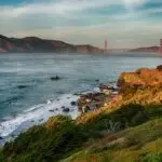 A coastal landscape with the Golden Gate Bridge in the background, partially covered by fog. Rocky cliffs and greenery line the shore, extending into the distance where rugged hills rise under a blue sky with scattered clouds. Waves gently crash against the shoreline—an ideal setting for one of Five Ways to Celebrate July 4th in the Bay Area.