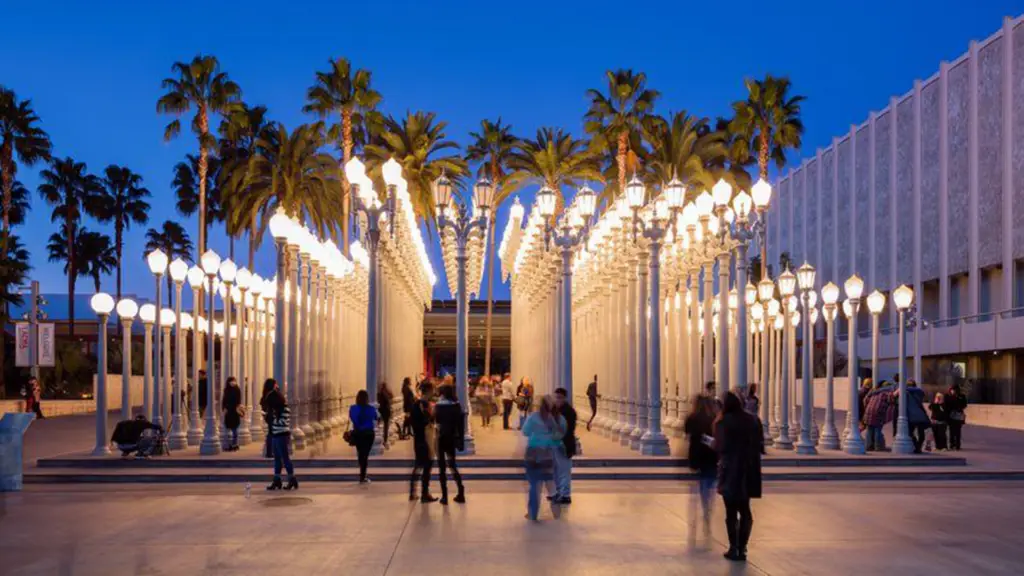 A group of people walk and gather among the tall, illuminated street lamps of the "Urban Light" installation at LACMA during early evening, exploring one of the best places to visit in Los Angeles, with palm trees and the museum building in the background.