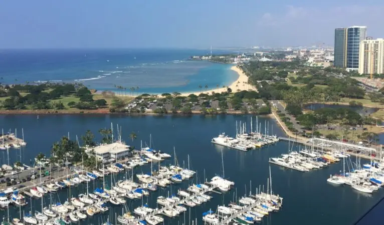 Aerial view of a marina filled with numerous boats docked in clear, blue water. Beyond the marina lies Magic Island Beach Oahu, sandy shores, lush green parks, and high-rise buildings. The ocean extends into the horizon, while the sky is clear and bright.