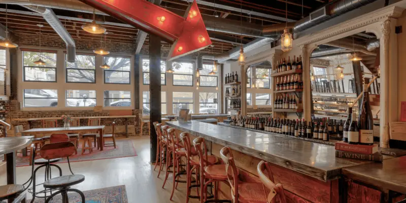 Cozy wine bar interior with rustic decor. A long bar counter lined with wooden stools, shelves filled with wine bottles, and hanging pendant lights create a warm ambiance. Large windows allow natural light to fill the space, and a red arrow sign adds a quirky touch—ideal for those exploring where to taste wine in San Francisco.