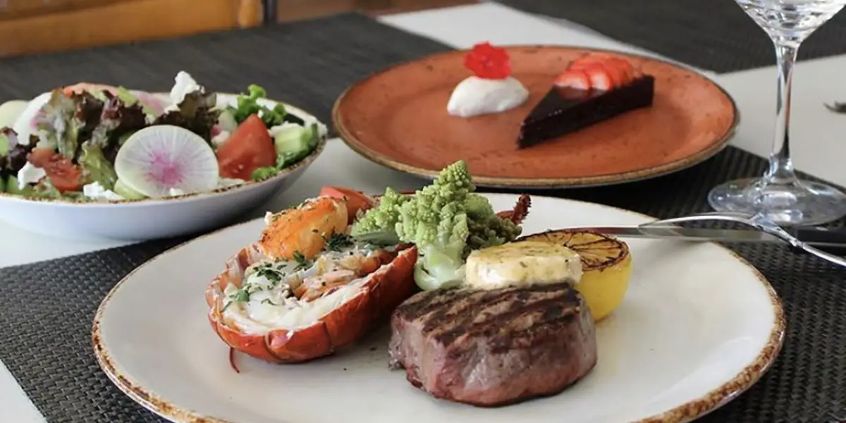 A gourmet meal is presented, featuring the best dinner on the Big Island: a surf-and-turf plate with grilled steak, lobster, and vegetables. A side salad with mixed greens and radish slices is also visible, along with a dessert plate showcasing a slice of chocolate cake and a dollop of cream.