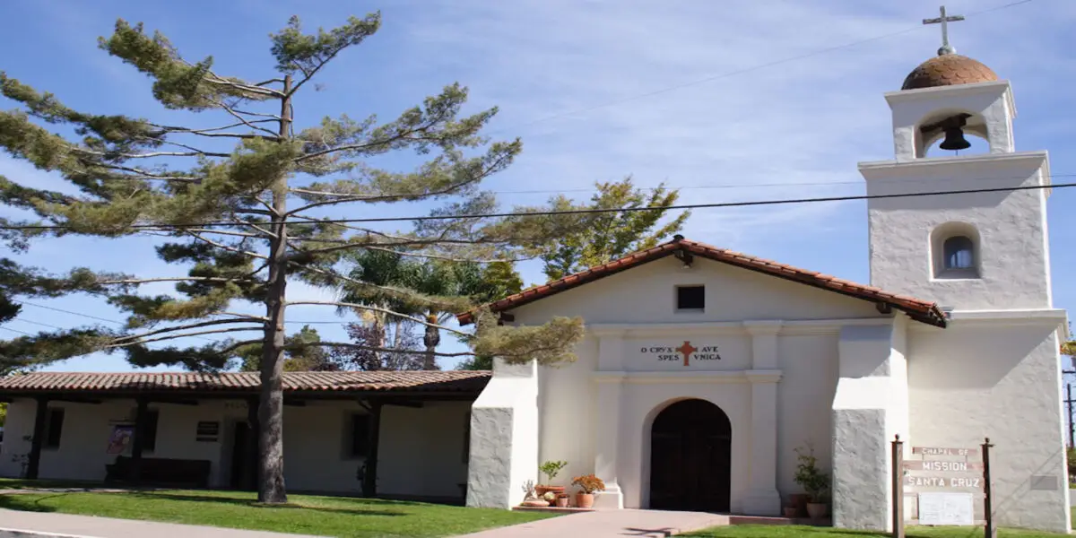 A small white mission-style church with a bell tower topped by a cross. The front entrance is an arched wooden door. Nearby is a tall tree and a signpost that reads "Historic Mission Santa Cruz," one of the iconic Northern California Missions. The sky is clear and blue.