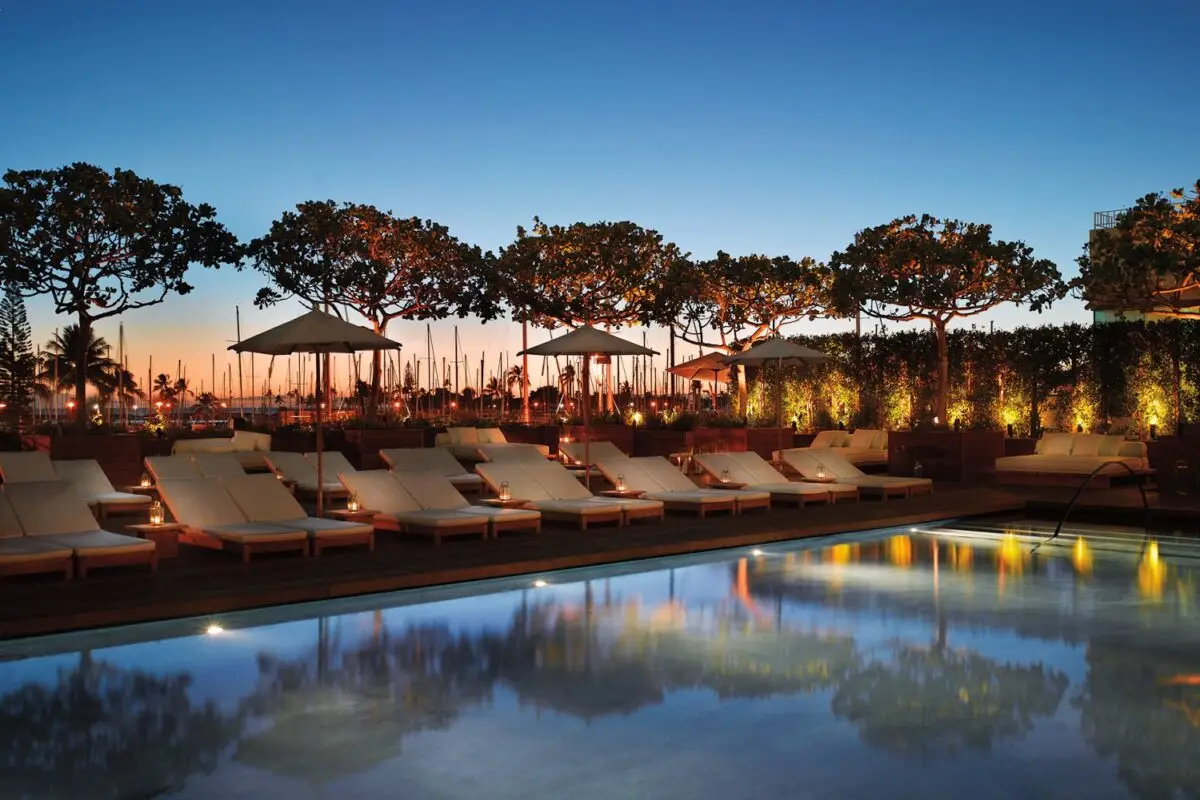 A serene outdoor pool area at dusk, featuring a row of empty lounge chairs and umbrellas lined along the poolside. Gently illuminated trees and soft lighting create a calm and inviting atmosphere, perfect for group getaways in Oahu, with reflections beautifully mirrored on the still water.