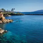 A picturesque coastal scene shows a house perched on a rocky cliff, overlooking clear blue waters. Small waves lap against the rocks, and a distant island is visible under a clear, blue sky. Scattered palm trees add to the tropical ambiance, reminiscent of the best romantic hotels in Maui.