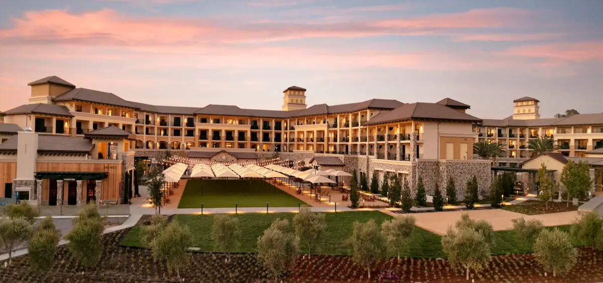 A large, elegant resort building with multiple levels and balconies, surrounded by well-manicured gardens and green lawns. The structure features tan walls, stones accents, and an impressive central courtyard. As the sky is painted with hues of pink and orange from the setting sun, you'll discover one of the best things to do in Napa Valley in November.