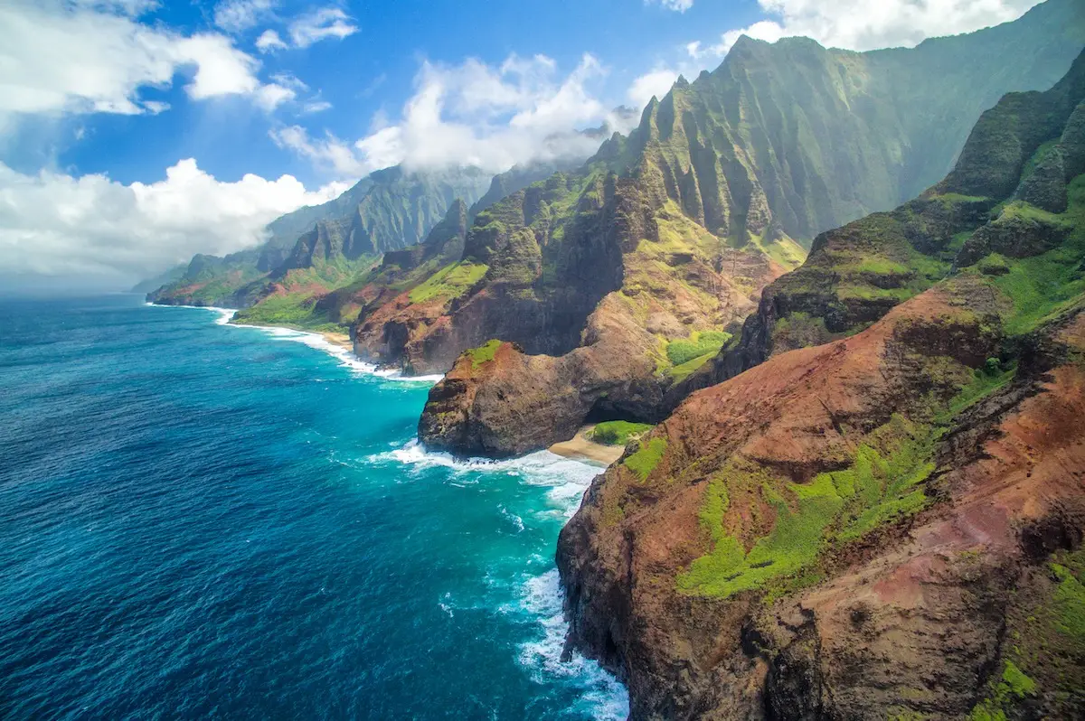 Aerial view of a rugged coastline with towering green cliffs, vibrant blue ocean waves crashing against the rocky shore, and patches of sandy beach. The sky is partly cloudy, with white clouds hovering over the lush landscape that evokes the spirit of Malama Kaua'i.