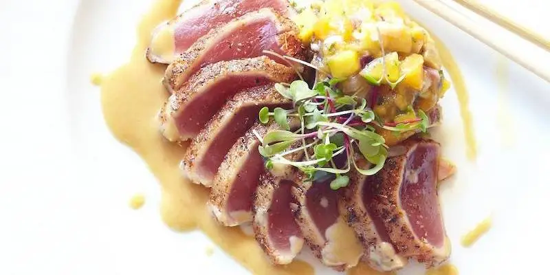 Sliced seared ahi tuna drizzled with a creamy sauce, garnished with microgreens and a mango salsa, plated on a white dish with chopsticks on the side. Experience Wailea's best gluten-free delight in every bite.