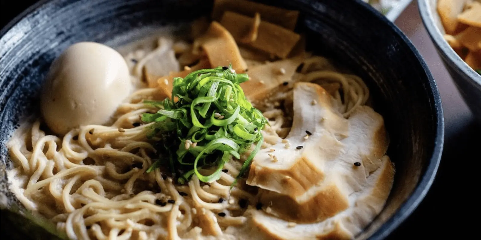 A close-up of a bowl of ramen found among the best cheap food Oahu offers. The dish includes curly noodles, slices of pork, bamboo shoots, a boiled egg, and garnished with chopped green onions. The creamy broth is sprinkled with sesame seeds.