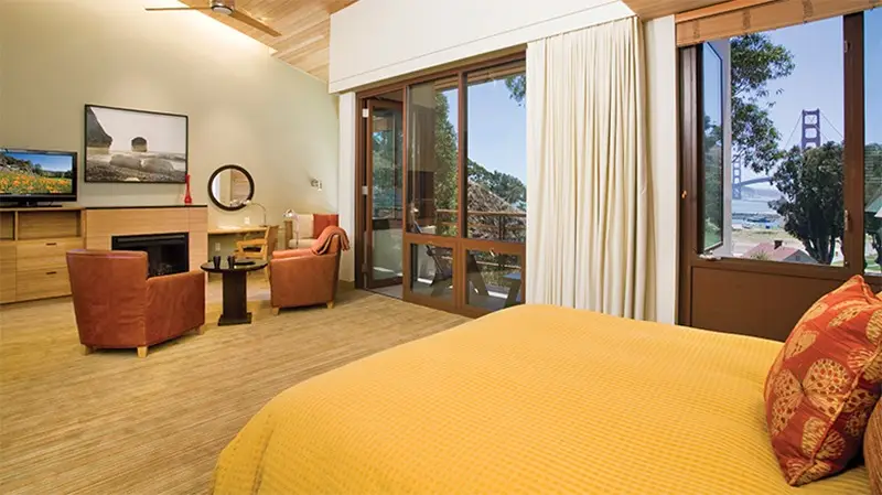 A cozy bedroom has a large bed with a yellow bedspread and vibrant pillows, a seating area with two chairs, a TV, a vanity desk, and a modern fireplace. The room features large windows and a balcony offering views of lush greenery and the Golden Gate Bridge, ideal for those staying at top Marin County hotels.