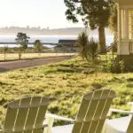 Four white Adirondack chairs face a serene view of a grassy field, a calm body of water, and the distant silhouette of a city skyline. Nearby, there's a charming house with a large porch, bathed in the warm glow of the setting or rising sun—a scene reminiscent of the tranquility offered by Marin County hotels.